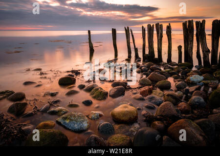 Alte Pole der Steg am steinigen Strand in warmen Orange und Sunrise, Behrensdorf, Ostsee, Norddeutschland Stockfoto