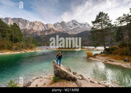 Junge Frau steht auf einem Felsen am Ufer, Blick in die Ferne, Eibsee vor Zugspitzmassiv mit Zugspitze, Wettersteingebirge, in der Nähe von Stockfoto