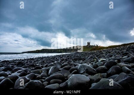 Schwarze, runde Felsen an der Küste, Dunstanburgh Castle mit bewölktem Himmel im Rücken, Craster, Northumberland, Großbritannien Stockfoto