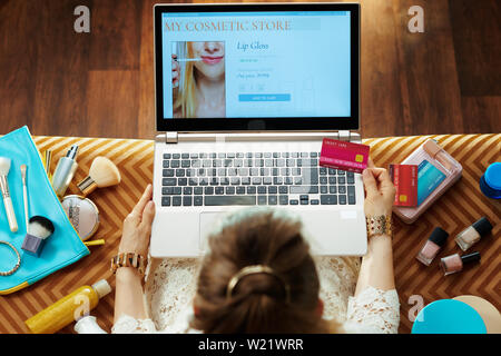 Obere Ansicht von eleganten Hausfrau in weißem Kleid mit Kreditkarte, Beauty Makeup online shopping auf einem Laptop beim Sitzen auf der Couch in der modernen Stockfoto