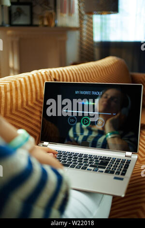 Entspannt junge Frau die Musik mit Kopfhörern auf einem Laptop auf der Couch in der modernen Haus im sonnigen heißen Sommertagen sitzen. Stockfoto