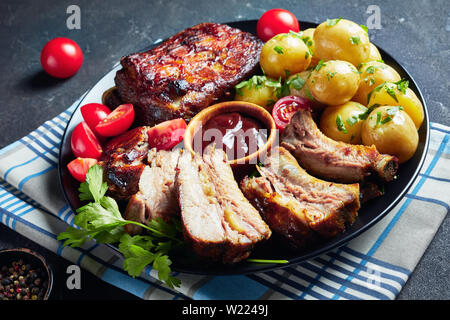 Close-up von gegrillten Schweinerippchen mit Barbecue Sauce mit Tomaten Salat serviert und heißes neuen Kartoffeln auf einer schwarzen Platte auf einer konkreten ta serviert. Stockfoto