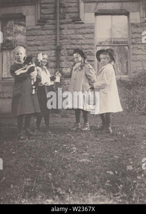 Jahrgang des frühen 20. Jahrhunderts photographische Postkarte zeigt eine Gruppe von drei britischen Schule Alter Mädchen Spaß zusammen haben. Eines der Mädchen hält einen Hund. Einer hält einen Korb. Die anderen beiden sind mit einer Tasse und einem Krug. Stockfoto