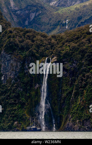 Einen Milford Sound Cruise Boot unter einem Wasserfall, Fiordland National Park, South Island, Neuseeland Stockfoto