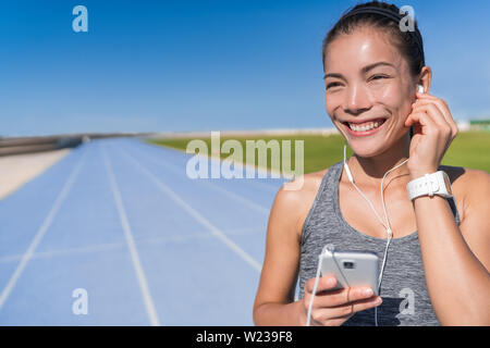 Asiatische runner Frau hören, die motivation Musik mit Ohrhörern auf Ihrem Handy app für Laufen auf der Schiene und im Feld Blue Lanes Titel in der Leichtathletik Stadion. Athleten genießen Fitness aktiv leben. Stockfoto