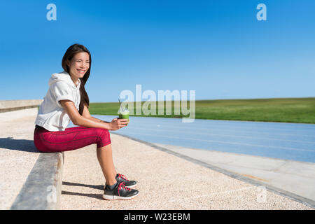 Gesundes Leben runner Sportler trinken grünen Smoothie frühstück Saft trinken vor dem Rennen für einen aktiven Lebensstil. Asiatische fitness laufen Frau glücklich bei Stadion blau Titel vor cardio Workout suchen. Stockfoto