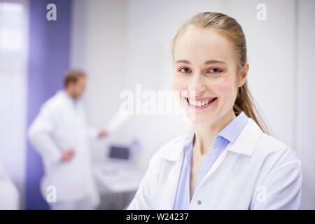 Ärztin lächelnd in der Klinik, männlicher Arzt im Hintergrund. Stockfoto