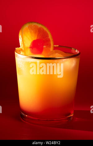 Tequila Sunrise in ein Glas mit goldenen Rand garniert mit Orange Slice und Kirsche. Roter Hintergrund, hohe Auflösung Stockfoto