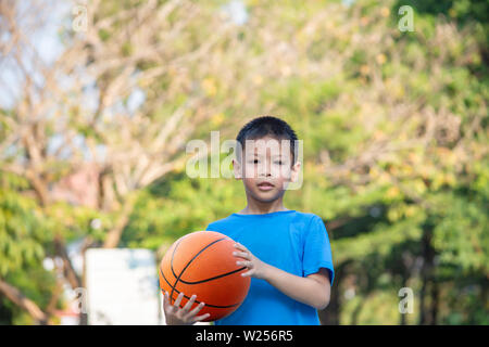 Asian Boy Holding ein Basketball Ball Hintergrund verschwommen Bäume. Stockfoto