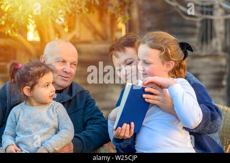 Glückliche Großeltern genießen Ihre Grand Kinder außerhalb. Family Portrait. Schöne Glückliche alte Paar Enkelinnen Buch zusammen Lesen in der Natur bei Sonnenuntergang. Stockfoto