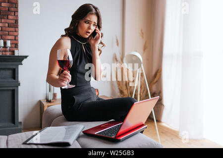 Junge Geschäftsfrau, die zu Hause arbeiten. Das Sitzen am Tisch und am Telefon sprechen. Glas Rotwein in der Hand halten. Brunette Modell tragen schwarze und braune sha Stockfoto