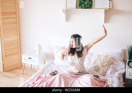 Dunkelhaarige schöne junge brunette, in ihrem Bett. Ahnd Strecken und Gähnen. Allein im Schlafzimmer. Schlafbrille auf der Stirn. Bis in den Morgen Stockfoto