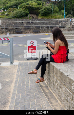 Junge Frau in Rot und Schwarz tragen mit einem Smartphone während draußen sitzen auf einer Mauer. Stockfoto