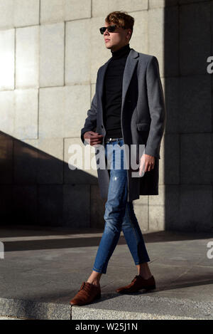 Modische junger erwachsener Mann in der Sonnenbrille. Posieren in Grau elegante Jacke, schwarz Rollkragen, Jeans und Braune mocassin Schuhe. Modetrends. Stockfoto