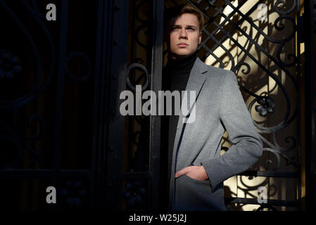 Stilvolle Mann im schwarzen Rollkragenpullover und Melange Grau Jacke, stehend in der dekorativen Metal Gate, ein bisschen in der Sonne und Schatten. Stockfoto