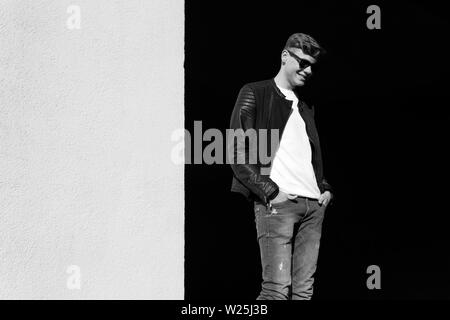 Lächelnd stilvolle junger Mann, stehend auf schwarzen und weißen Hintergrund. Männliche Mode - Lederjacke, Jeans, weißes Hemd und Sonnenbrille. Stockfoto