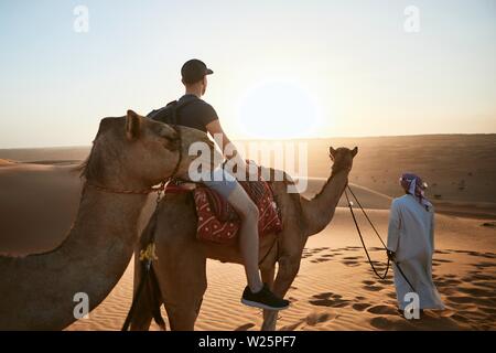 Kamelreiten in der Wüste bei Sonnenuntergang. Junger Mann genießen Reise auf Sanddünen. Wahiba Sands im Sultanat Oman Stockfoto