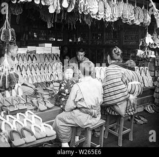 [1900s Japan - Geta schuhe Shop] - Kunden in einem Geschäft mit geta (holzschuhe) und waraji (Stroh Sandalen). 20. Jahrhundert vintage Glas schieben. Stockfoto