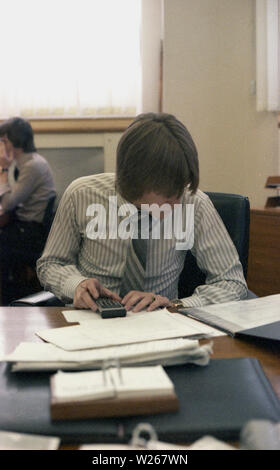 1970 s, historischen, einem jungen erwachsenen männlichen, Ende zwanzig oder Anfang dreißig im Alter in einem Büro am Schreibtisch sitzen, arbeiten, eine finanzielle Manager oder möglicherweise ein Buchhalter, wie er ist, mit einem kleinen Taschenrechner Zahlen von gedruckten Büchern oder Zeitschriften, England, UK. Stockfoto