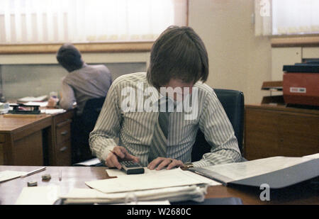 1970 s, historischen, männlich, Ende zwanzig oder Anfang dreißig im Alter in einem Büro am Schreibtisch sitzen, arbeiten, eine finanzielle Manager oder möglicherweise ein Buchhalter, wie er ist, mit einem kleinen Taschenrechner Zahlen von gedruckten Büchern oder Zeitschriften, England, UK. Stockfoto