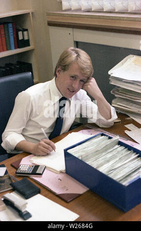 1970 s, historischen, einem jungen erwachsenen männlichen, Ende zwanzig oder Anfang dreißig im Alter in einem Büro am Schreibtisch sitzen, arbeiten, eine finanzielle Manager oder möglicherweise ein Buchhalter, wie er mit einem Taschenrechner Zahlen von gedruckten Büchern oder Zeitschriften, England, UK. Stockfoto