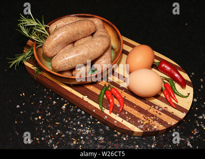 Frische Würstchen in einer Schüssel auf einem Holzbrett mit Eier und rote Chilischoten Stockfoto