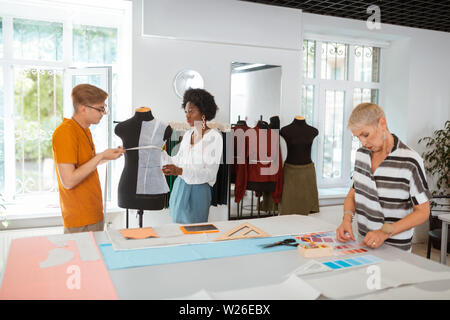 Kreatives Team. Zwei junge Modedesigner von Messungen an der Schneiderpuppe, während ihre älteren Mitarbeiter Auswahl der Farbpalette Stockfoto