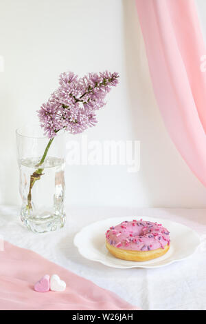 Moderne Stillleben mit lila Zweig, Donut auf dar, Platte, rosa Stoff und kleinen herzförmigen Marshmallows auf weißem Hintergrund. Stockfoto