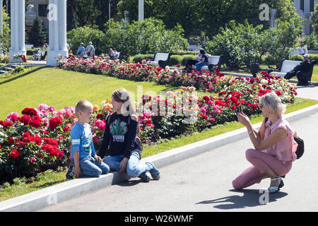 Moskau, Russland - Juli 2, 2019, Mutter nimmt Bilder ihrer Kinder auf dem Hintergrund einer Bett von Rosen Stockfoto