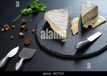 Keile von semi-soft Käse auf einem schwarzen Holz- Käseplatte mit Stahl Cutter und zerstreute gehackte Walnüsse in einem hohen Winkel food Still Life Stockfoto