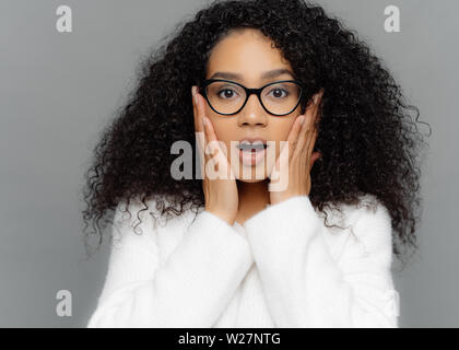 Close up Portrait von schockiert betroffene junge Frau mit dunkler Haut und lockiges Haar, hört erschreckende Nachrichten, hält die Hände auf die Wangen, trägt eine Brille und w Stockfoto