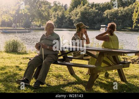 Zwei älteren Damen und ein alter Mann sitzt auf einer Picknickbank, die Frauen Interaktion, der ältere Mann hat einen Stock, und er ist das Ignorieren der Damen. Stockfoto