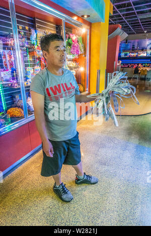 Ein im mittleren Alter Asiatischen männlich, in Shorts und T-Shirt gekleidet, steht in einem kinder Arcade mit einer Faust voller gewinnende Karten wie er schaut über die Preise Stockfoto