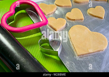 Zwei herzförmige Ausstechformen in einem Cookie Backblech; Schnitt Cookies im Hintergrund. Stockfoto