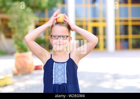 Happy girl In Gläsern mit Apfel auf den Kopf. Große Porträt der Schüler außerhalb des Unterrichts. Zurück zu Schule, Balance, Erfolg, Ziel und gesundes Essen für Kinder Konzept. Stockfoto