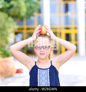 Happy girl In Gläsern mit Apfel auf den Kopf. Große Porträt der Schüler außerhalb des Unterrichts. Zurück zu Schule, Balance, Erfolg, Ziel und gesundes Essen für Kinder Konzept. Stockfoto