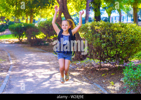 Kleines Mädchen auf der sonnigen Park in der Tageszeit springen. Glücklich lächelnde Kind zurück in die Schule. Schüler mit Tasche klicken Sie auf der Grundschule. Volle Länge Portrait von fröhlichen Kind springen outdoor Herbst Hintergrund. Stockfoto