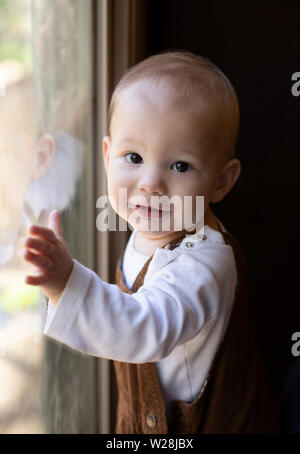 Kleinkind/Baby in Overalls stehen am Fenster, mit Blick auf die Kamera Stockfoto