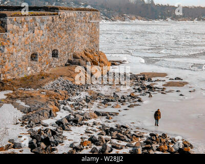 Ein einsamer Mann Spaziergänge entlang der eingefroren und felsigen Strand außerhalb der Mauern von Suomenlinna Festung auf einer Insel direkt vor der Küste von Helsinki, Finnland. Stockfoto