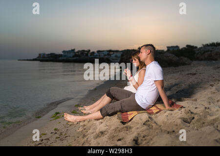 Junge romantische Paar entspannen am Strand zu sitzen und die gemeinsame Nutzung von Wassermelone am Abend Stockfoto
