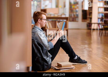 Clevere Kerl in Denim Hemd und schwarzen Jeans lesen durch Bücherregal Stockfoto