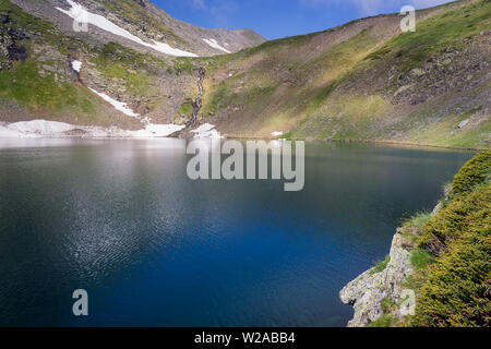 Vordergrund sonnenbeschienenen Juniper, Deep Blue das Auge Lake im Rila Gebirge, malerischen Wasserfall die Kaskadierung der felsigen Klippen und fernen Gipfel Stockfoto