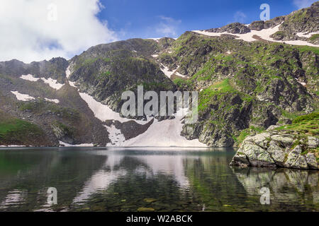 Grüne Gras auf einer felsigen Hochland und Spitzen von Rila Gebirge und Reflexionen im sauberen Wasser des Auges Gletschersee, einer der Sieben Rila-Seen Stockfoto