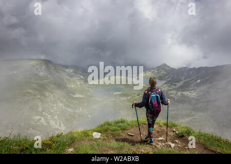 Ein Mädchen mit bunten Strumpfhosen, Rucksack und Wanderstöcke stehen am Rand des Sees Peak auf Rila-gebirge, berühmten Aussichtspunkt von Sieben Rila-Seen Stockfoto