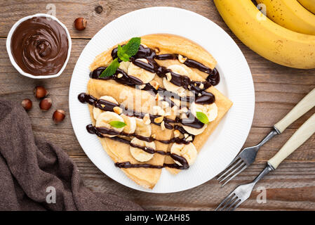 Pfannkuchen mit Banane und Schokosauce und Haselnuss Schokoladencreme auf weiße Platte garniert mit gehackten Nüssen. Rustikalen Holztisch Hintergrund, Tabelle Stockfoto