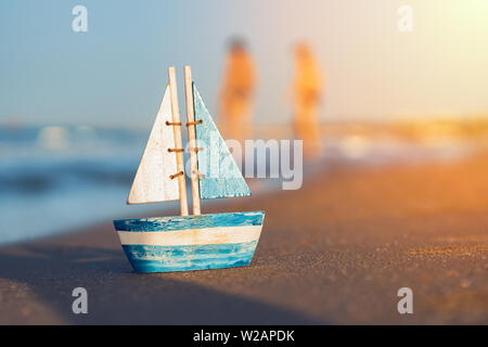 Holzspielzeug Segelboot auf Sand in der Nähe der Küste im Sommer. Verschwommene Menschen und Spielzeug Segelboot am Strand. Urlaub und Freizeit Konzept Konzept. Stockfoto