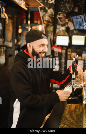 Lächelnd Barkeeper Bier in einer Bar. Bärtige mutiger Mann gießt sie einem Schäumenden Getränk. Goldener Hahn für Bier. Stockfoto