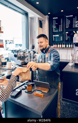 Lächelnd bärtigen kaukasischen Mann Kassierer Barista im Restaurant Cafe bei der Arbeit. Verkäufer geben Client Kunden ihren Kauf Essen in Brown Organic Papier packa Stockfoto