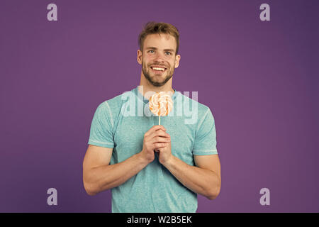 Mann essen großen bunten süßen Lutscher. Mann mit Borsten mag Lollipop. Cheat meal Konzept. Zucker schädlich für die Gesundheit. Kerl halten Lollipop candy violett unterlegt. Geschmack der Kindheit. Lollipop Spaß. Stockfoto