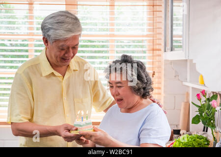 Asiatische älterer Mann überraschen ältere Frau mit Geburtstag Kuchen in der Küche zu Hause. Das Altern zu Hause Konzept Stockfoto